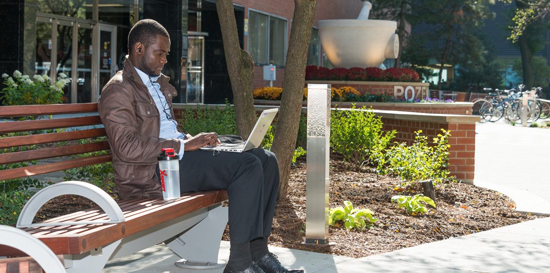 a man sits on a bench with his laptop next to POZEN Plaza and the UIC Pharmacy Wood Street building