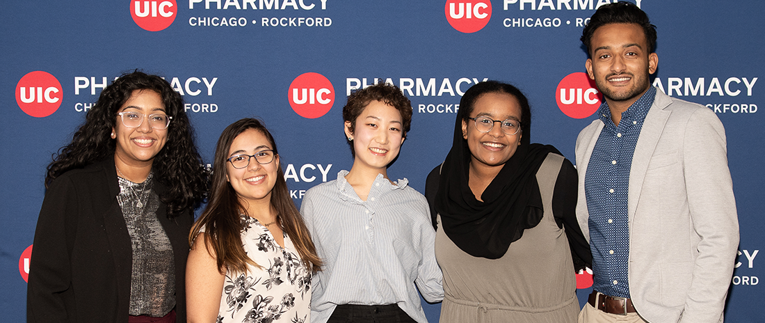 5 Pharmacy students line up for a photo in front of a UIC Pharmacy logo background