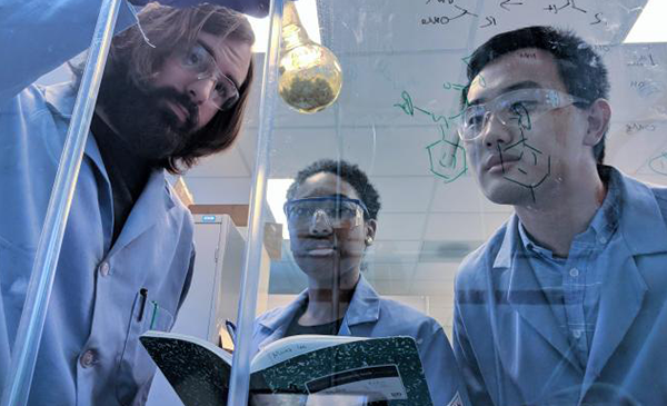 Photo of three scientists standing in front of a fume hood; one is holding a flask, and another is holding a lab notebook