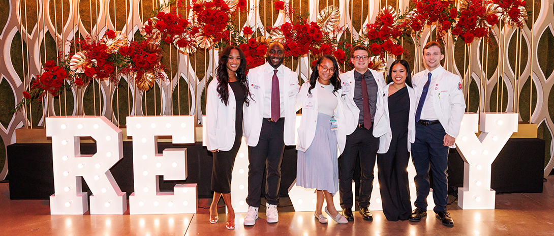 5 Pharmacy students line up for a photo in front of a UIC Pharmacy logo background
